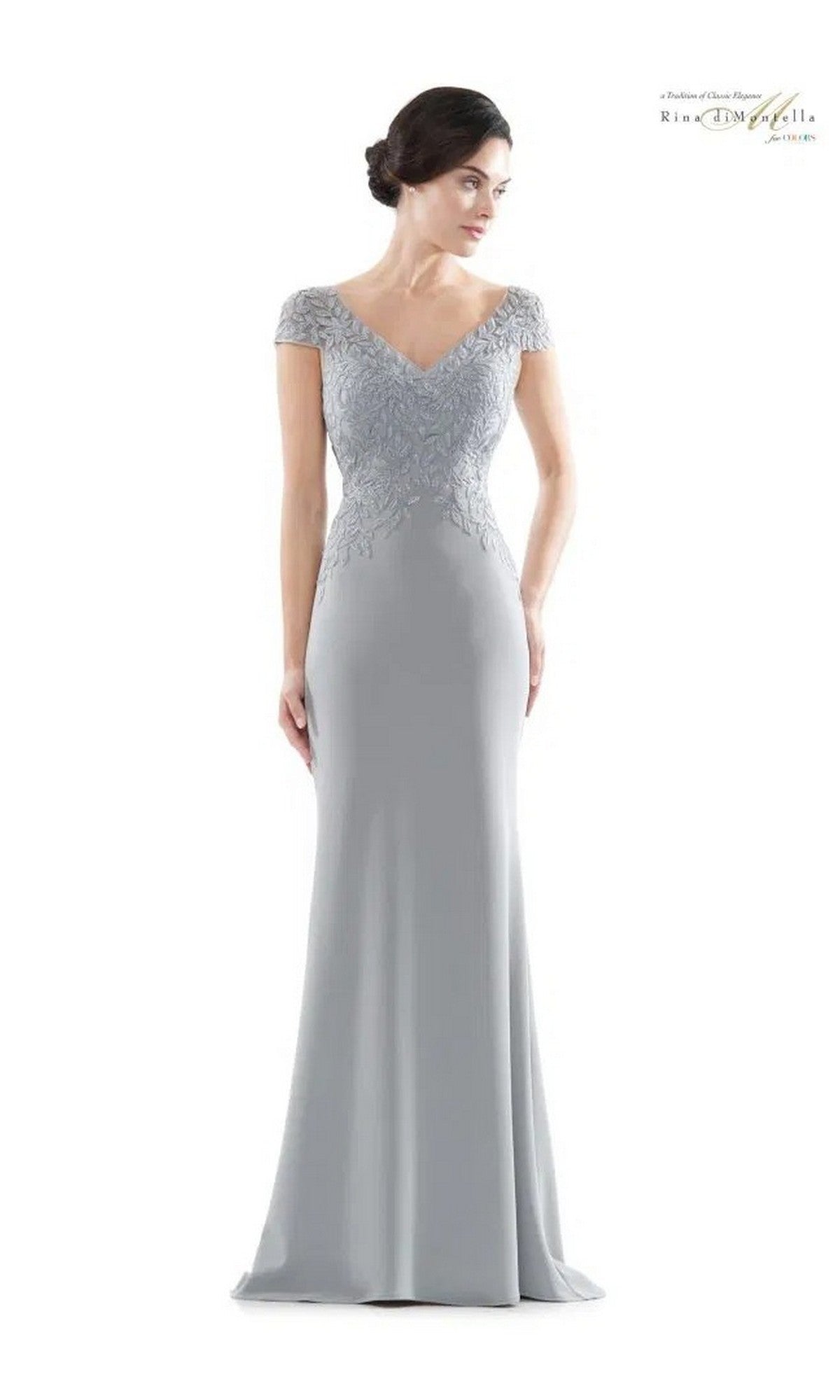 A woman models the Rina Di Montella RD2718, a long formal dress in light gray with short sleeves, an embroidered bodice, and a V-neckline, standing against a plain white background.